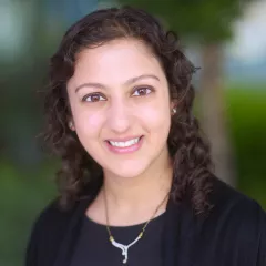 Headshot of a smiling woman with light skin tone and wavy dark hair wearing a dark top against a blurred outdoor background