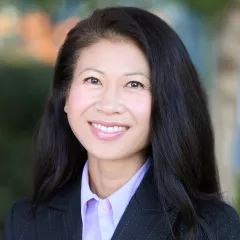 Headshot of a smiling woman with light skin tone and straight dark hair wearing a purple blouse under a dark suit jacket against a blurred outdoor background