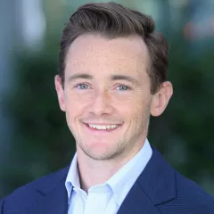 Headshot of a smiling man with light skin tone and brown hair wearing a light blue dress shirt and dark blue suit jacket against a blurred outdoor background