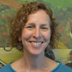 Headshot of a smiling woman with light skin tone and short, curly, auburn-colored hair wearing gold ringed earrings and a light blue sweater against a colorful painting