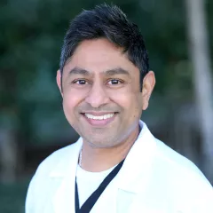 Headshot of a smiling man with medium skin tone and short dark hair wearing a white lab coat against a blurred outdoor background