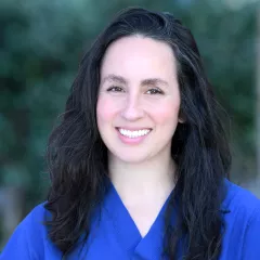 Headshot of a smiling woman with light skin tone and straight dark hair wearing blue surgical scrubs against a blurred outdoor background