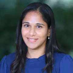 Headshot of a smiling young woman with medium skin tone and dark hair wearing a blue top against a blurred outdoor background