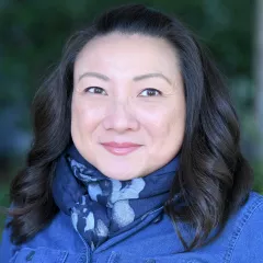 Headshot of a smiling woman with dark, shoulder-length hair wearing a blue scarf and blue denim shirt against a blurred outdoor background