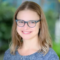 Headshot of a smiling woman with light skin tone and shoulder-length brown hair, wearing grey framed glasses, gold hoop earrings and a grey sweater against a blurred outdoor background