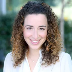 Headshot of a smiling woman with light skin tone and curly, shoulder-length brown hair wearing a white blouse against a blurred outdoor background