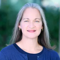 Headshot of a smiling woman with light skin tone and shoulder-length grey hair wearing small gold hoop earrings and a dark blue top under a blue cardigan against a blurred outdoor background