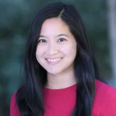 Headshot of a smiling woman with light skin tone and long dark hair wearing a fuchsia top against a blurred outdoor background