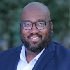 Headshot of smiling man with dark skin tone, a shaven head and a dark beard wearing black-rimmed eyeglasses and an open-collar light blue dress shirt under a dark blue suit jacket against a blurred outdoor background