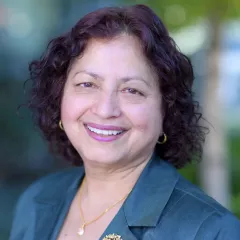 Headshot of a smiling woman with medium skin tone and short dark wavy hair wearing small gold hoop earrings, a gold necklace and green suit jacket against a blurred outdoor background