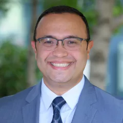 Headshot of a smiling man with medium skin tone and short dark hair wearing black framed eyeglasses, a light dress shirt and dark blue tie under a blue suit jacket against a blurred outdoor background