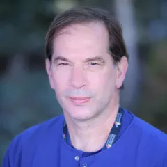 Headshot of a man with light skin tone and brown hair wearing a blue round-collar shirt against a blurred outdoor background