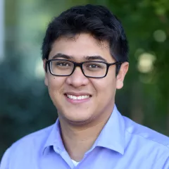 Headshot of a smiling man with medium skin tone and dark hair, wearing black eyeglasses and open-collar purple dress shirt against a blurred outdoor background