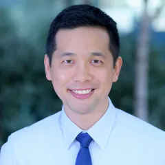 Headshot of a smiling man with medium skin tone and short dark hair wearing a light blue dress shirt and solid blue tie against a blurred outdoor background