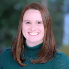 Headshot of a smiling woman with light skin tone and shoulder-length auburn hair wearing a green turtleneck sweater against a blurred outdoor background