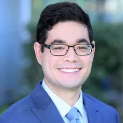 Headshot of a smiling man with light skin tone and dark hair wearing black-framed eyeglasses, a light blue dress shirt and blue tie under a blue suit jacket against a blurred outdoor background