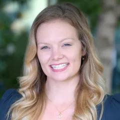 Headshot of a smiling woman with light skin tone and long blonde hair wearing a small gold necklace and dark blue top against a blurred outdoor background