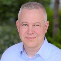 Headshot of a man with light skin tone and short grey hair wearing a blue, open-collar dress shirt against a blurred outdoor background