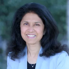 Headshot of a smiling woman with medium skin tone and shoulder-length dark hair wearing a black top under a light blue suit jacket against a blurred outdoor background