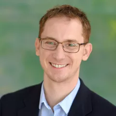 Headshot of a smiling man with light skin tone and auburn hair wearing rectangular eyeglasses and an open-collar light blue dress shirt under a dark suit jacket against a blurred outdoor background