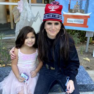 A young girl with medium skin tone wearing a pink dress sits with a woman with medium skin tone wearing a menorah beanie in front of a happy holidays poster