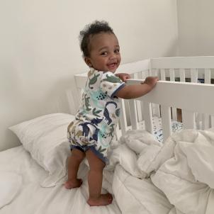 Standing infant with dark skin tone and dark hair wearing onesie with colorful dinosaur print smiles as they stand up supporting themselves on a crib railing