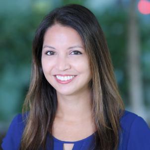 Headshot of a smiling woman with light skin tone and straight brown hair wearing a blue blouse with blurred outdoor background