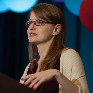 Woman with light skin tone and straight brown hair wearing black rimmed glasses and a tan top speaks behind lectern