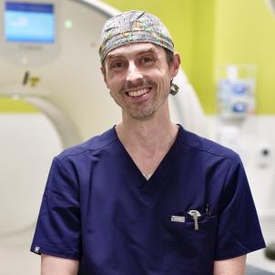 Male physician with light skin tone wearing purple scrubs and colorful surgical cap smiles with CT/fluoroscopy machine in the background