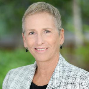 Headshot of smiling woman with light skin tone and short grey hair wearing dark teardrop earrings and a black top under a plaid grey suit jacket against a blurred outdoor background