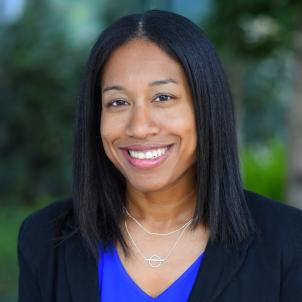 Headshot of a smiling woman with dark skin tone and dark shoulder-length hair wearing a blue top under a dark suit jacket