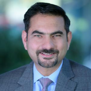 Headshot of a smiling man with medium skin tone, dark hair and a goatee wearing a blue dress shirt and purple tie under a dark grey suit jacket against a blurred outdoor background