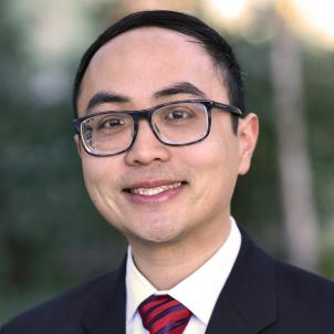 Headshot of a smiling man with light skin tone and dark hair wearing black rimmed glasses, white dress shirt and red and blue striped tie under a dark suit jacket against a blurred outdoor background