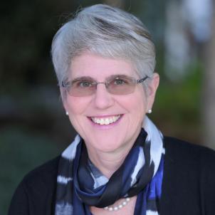 Headshot of a smiling woman with light skin tone and grey hair wearing tinted, rimless glasses, a patterned silk scarf and dark jacket against a blurred outdoor background