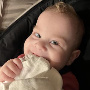 Male toddler with light skin tone and blonde hair smiles while chewing on their cream-colored blanket