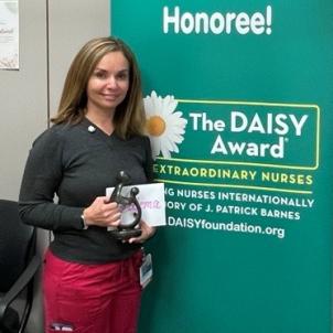 Smiling female nurse with medium skin tone and shoulder-length brown hair holds an award statue as they pose in front of congratulatory poster in a hospital setting