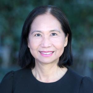 Headshot of smiling woman with light skin tone and shoulder-length dark hair wearing a black top against a blurred outdoor background