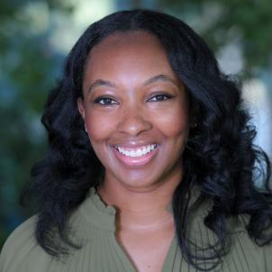 Headshot of a smiling woman with dark skin tone and wavy dark hair wearing a green top and small gold cross necklace against a blurred outdoor background