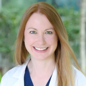 Headshot of a smiling woman with light skin tone and long, light brown hair wearing a blue top under a white lab coat against a blurred outdoor background