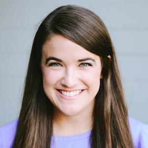 Headshot of a smiling woman with light skin tone and straight brown hair wearing a lavender top against a neutral indoor background