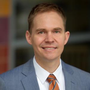 Headshot of a smiling man with light skin tone and short, light brown hair wearing a white dress shirt and orange and blue striped tie under a grey suit jacket against a blurred outdoor background