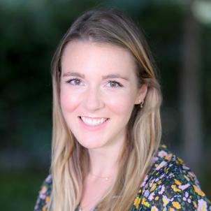 Headshot of a smiling woman with light skin tone and long blonde hair wearing small gold earrings and necklaces and a floral print dress against a blurred outdoor background