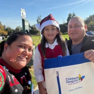 Young boy with medium skin tone and long brown hair wearing a Santa hat smiles as he poses with his mother and father on a bright, sunny day