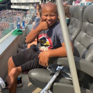 A medium-skinned boy sitting in a stadium smiles and flashes a thumbs up. A metal prosthesis is leaning against the seat.