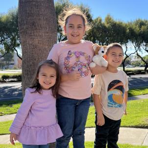 Two young girls and a young boy with medium skin tone and brown hair smile as they pose for a picture together on a bright sunny day