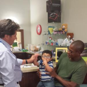 A toddler with glasses sits on his father’s knee, while a man adjusts the glasses.