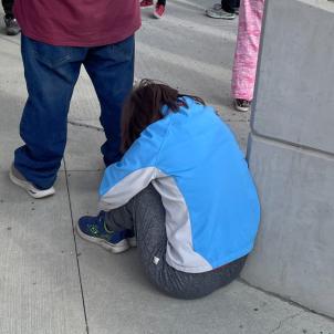 Medium shot of a light-skinned young adult woman sitting on the pavement and hunched over at an outdoor event