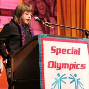 Medium shot of a light-skinned young adult woman standing on a stage at a podium with the Special Olympics logo on it and speaking into a microphone