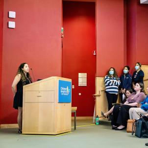 A young adult woman with medium-light skin tone stands at a lectern with the Children’s Hospital Los Angeles logo on it in front of a red wall in an auditorium, delivering a presentation to seated and standing audience members.