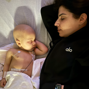 Small male child with bald and light skin sleeping in hospital bed next to woman with dark hair and light skin